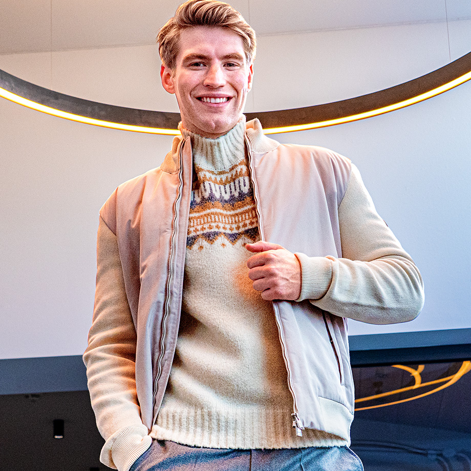 Lächelnder Mann in beigem Norwegerpullover und hellem Blouson unter modernem Lichtring – gemütliche Strickmode für stilvolle Winterlooks bei Herrenausstatter.de.