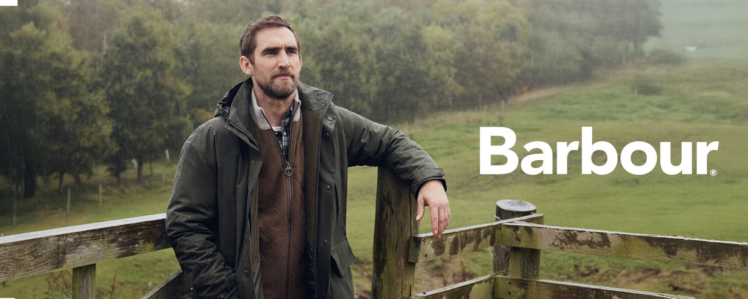 Mann in brauner Strickjacke, kariertem Hemd und grüner Outdoor-Jacke von Barbour lehnt an einem Holzzaun in ländlicher Landschaft.