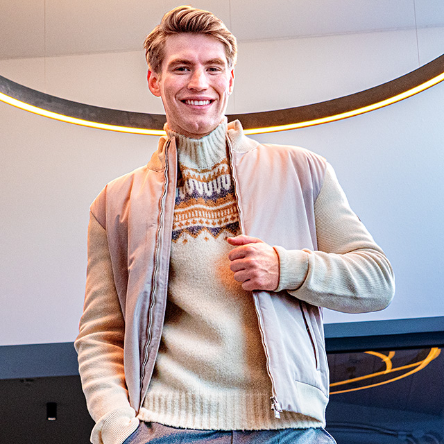 Lächelnder Mann in beigem Norwegerpullover und hellem Blouson unter modernem Lichtring – gemütliche Strickmode für stilvolle Winterlooks bei Herrenausstatter.de.