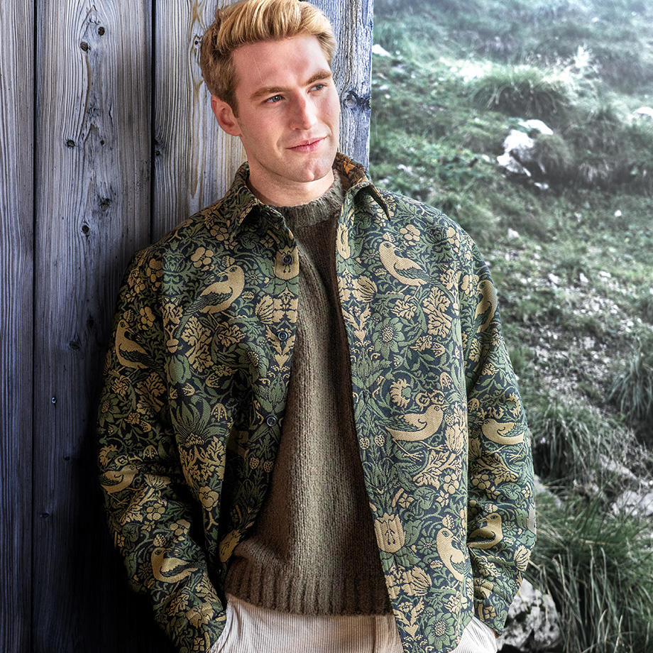 Mann trägt gemustertes Overshirt mit floralen Motiven und Strickpullover, steht vor Holzwand in alpiner Umgebung.