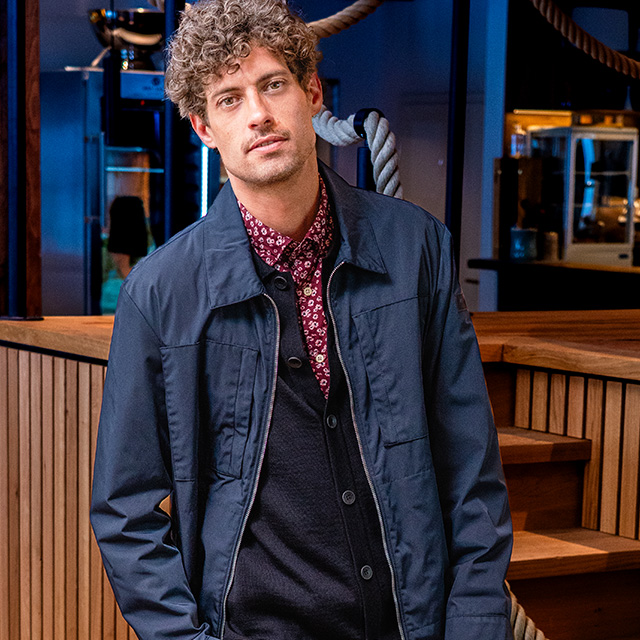 Mann in dunkelblauer Übergangsjacke, rotem gemusterten Hemd und dunkler Strickweste steht vor edler Holzeinrichtung – stilvolle Herrenmode mit modernem Twist entdecken bei Herrenausstatter.de.