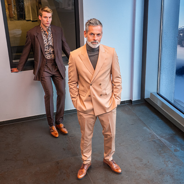 Zwei Männer in Anzügen stehen in modernem Loft, einer in Braun, der andere in Beige.
