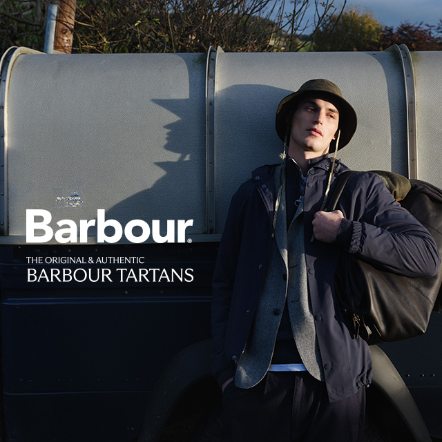 Mann in navyblauer Barbour-Jacke, grauem Schal und Fischerhut lehnt lässig an einem Land Rover – authentischer Outdoor-Look aus der Barbour Tartans Kollektion.
