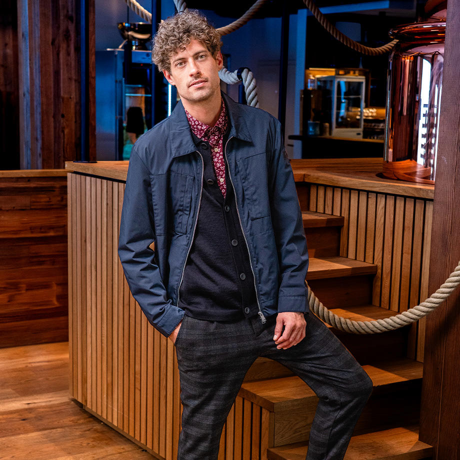 Mann mit lockigem Haar trägt ein legeres Outfit aus dunkelblauer Freizeitjacke, einem Hemd in der Trendfarbe Bordeaux und einer karierten Hose, fotografiert in moderner Location.