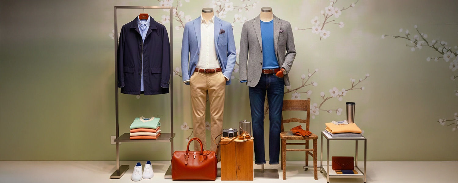Zwei Schaufensterfiguren in blauen und grauen Blazern, beige Chinos und Jeans, mit Accessoires vor floralem Hintergrund.