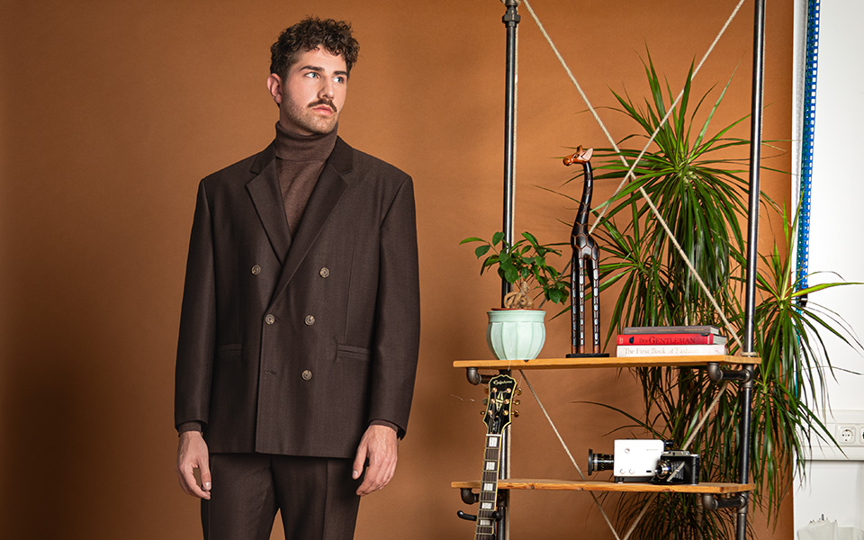 Mann in braunem Anzug mit Rollkragenpullover vor Regal