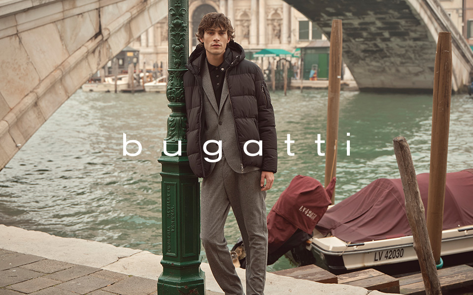 Mann in grauem Anzug mit schwarzer Daunenjacke, lässig am Flussufer lehnend