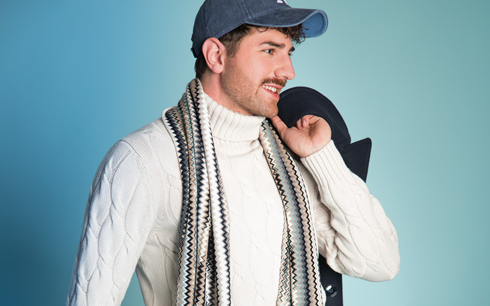 Weißer Strickpullover mit Zopfmuster, gemusterter Schal, Mann trägt blaue Kappe.