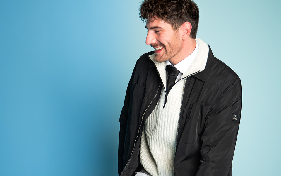 Mann in schwarzer Übergangsjacke über cremeweißem Pullover mit grauer Stoffhose, lässig sitzend