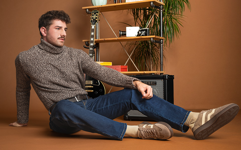 Herr in beiger Wolljacke und Jeans, sitzend mit Reisetasche, lässig präsentiert vor braunem Hintergrund