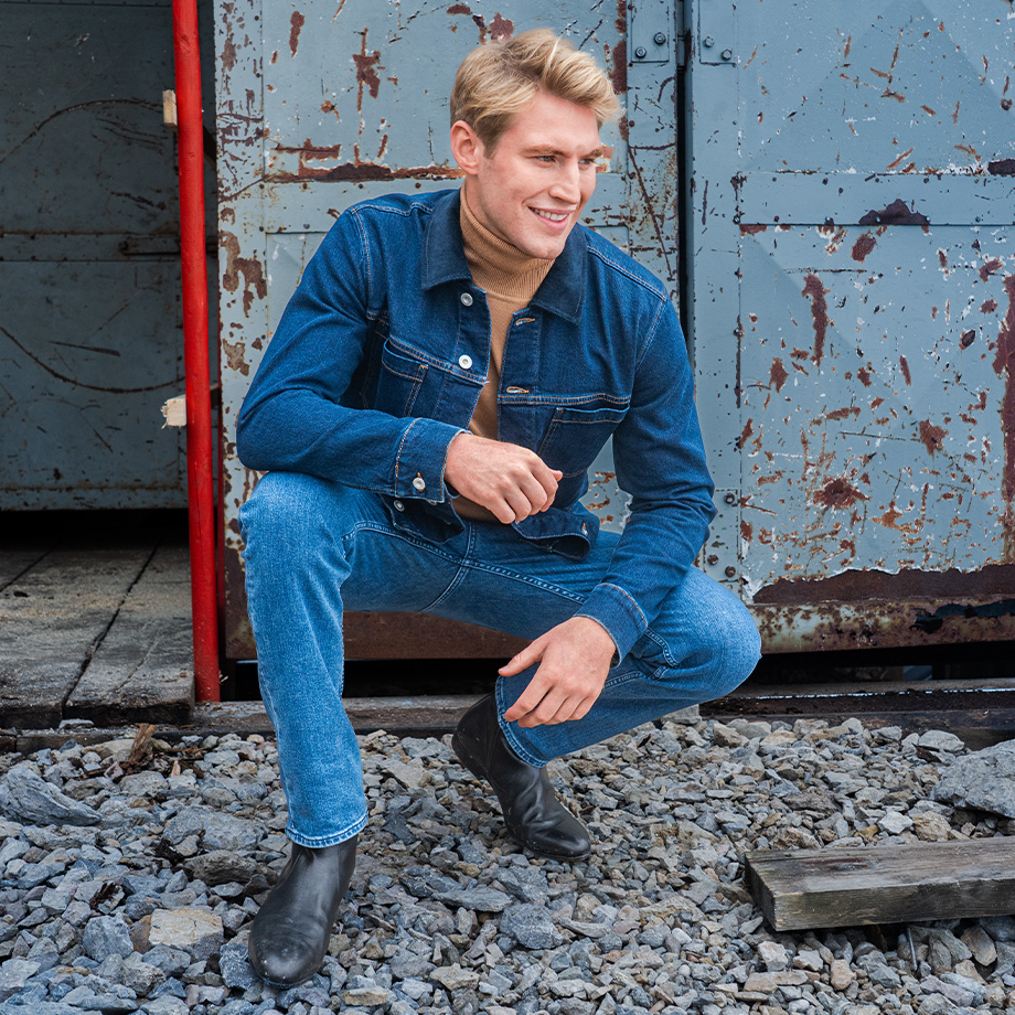 Mann in blauer Jeansjacke mit beigem Rollkragenpullover in lässiger Pose vor rostigem Tor