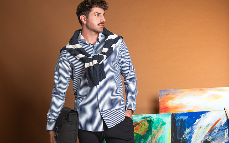 Mann in blau-weiß gestreiftem Hemd, Pullover über Schultern, schwarze Hose, mit Tasche, vor Gemälden