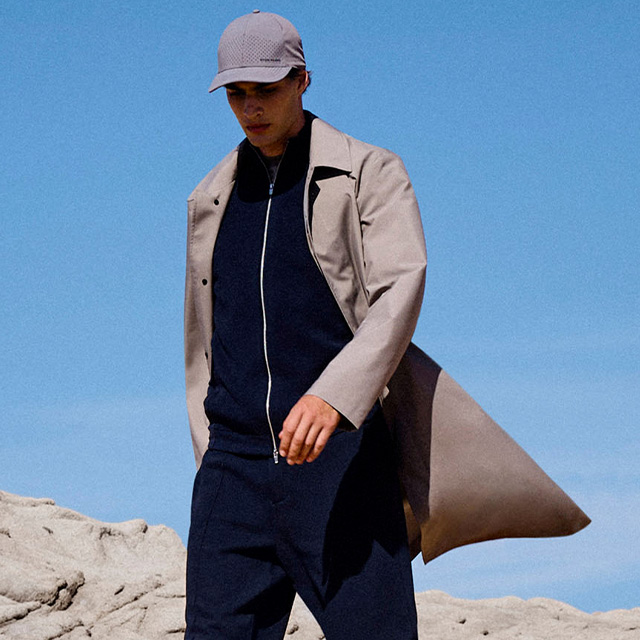 Beigefarbener Trenchcoat über dunkelblauem Reißverschluss-Sweatshirt, lässig getragen mit Kappe