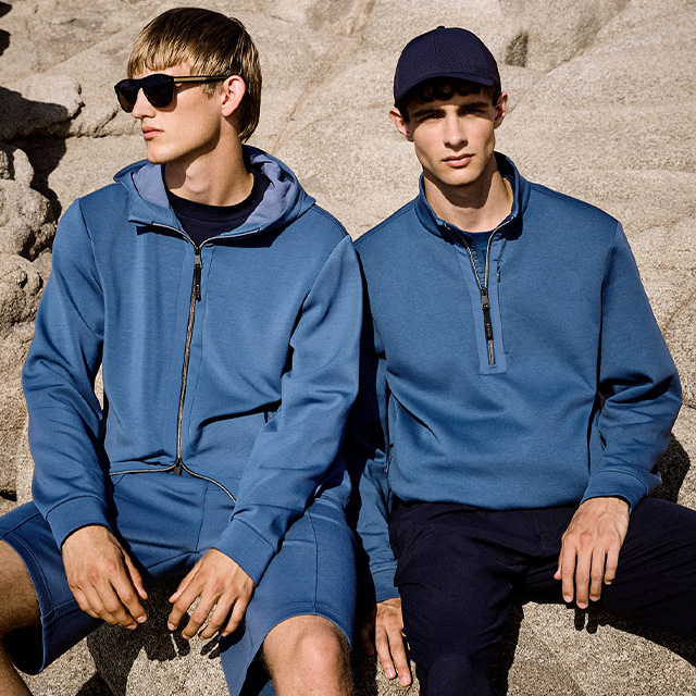 Zwei Männer in blauen Sweatshirts, einer mit Kapuzenjacke, im Freien sitzend.