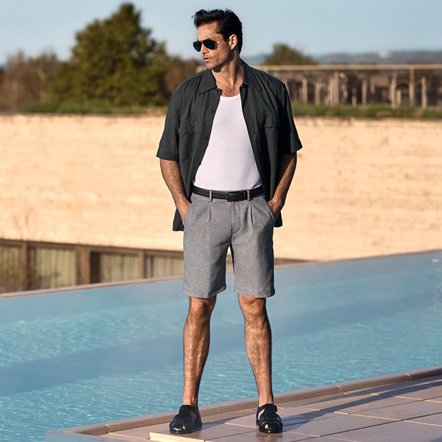 Mann in grauer Bermuda-Shorts und weißem Tanktop mit offenem Hemd am Pool