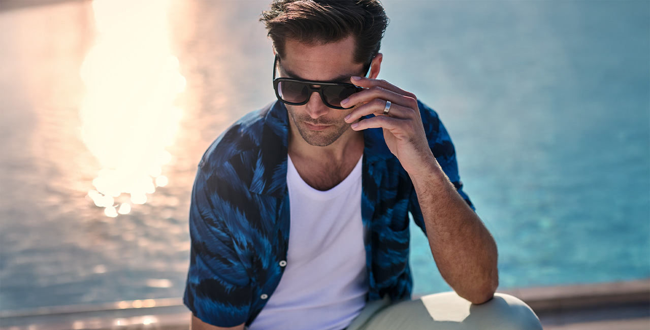 Mann in blauem Kurzarmhemd mit weißem T-Shirt am Wasser, trägt Sonnenbrille