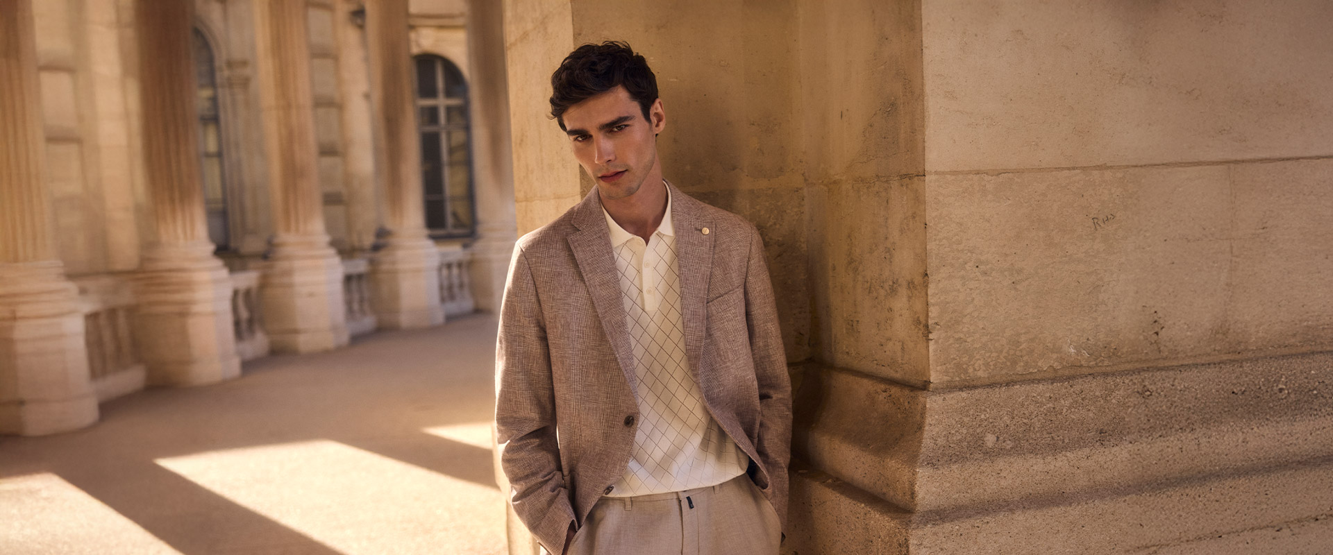 Mann in beigem Leinen-Sakko und cremefarbenem Polohemd mit Rautenmuster vor Säulen stehend