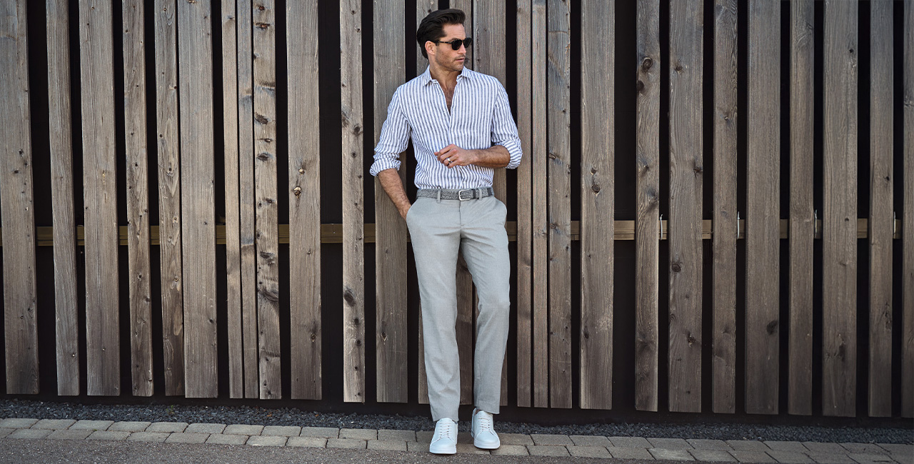 Herr in weiß-blau gestreiftem Hemd und grauer Hose lässig an Holzwand, weiße Sneaker, Freizeit-Look