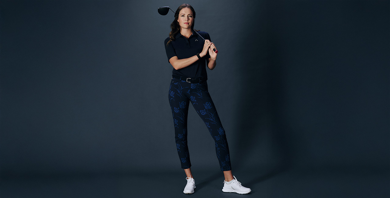 Eine Frau steht vor dunklem Hintergrund. Sie trägt eine dunkelbaue Golfhose mit blauem Blütenmuster dazu ein dunkelblaues Golfpolo.
