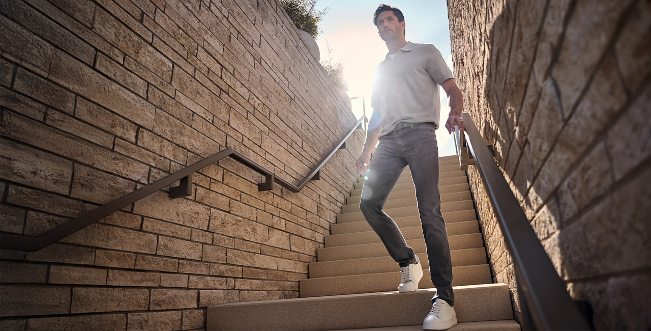 Grau meliertes Poloshirt und blaue Jeans, lässig auf Treppe getragen, Sonnenbrille in der Hand.