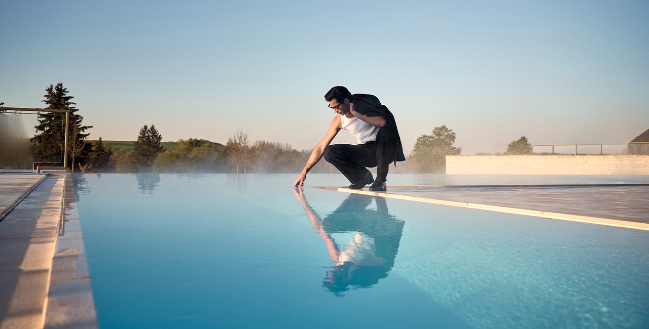 Mann in weißem Tanktop und schwarzer Hose am Rand eines Pools, Sonnenaufgang im Hintergrund