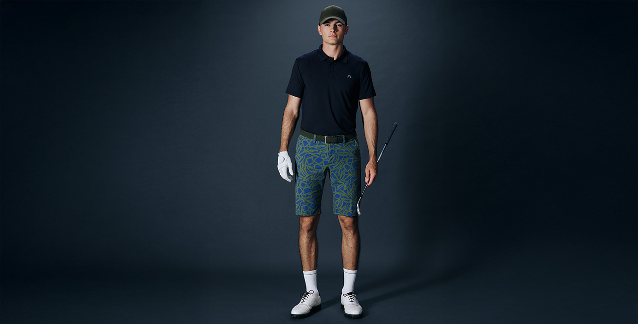 Herr in dunkelblauem Poloshirt und blau-grün gemusterten Golfshorts mit Schläger und Handschuh vor dunklem Hintergrund.