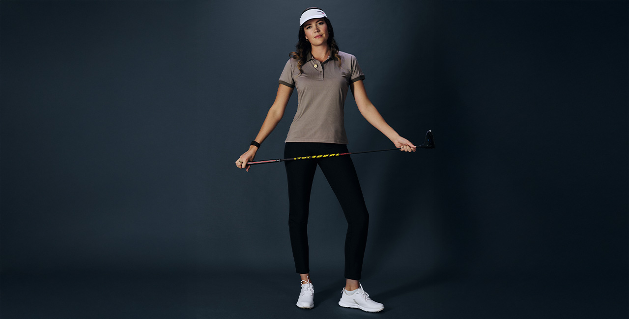 Eine Frau steht vor dunklem Hintergrund. Sie trägt eine schwarze Golfhose, dazu ein braunes Golfpolo und hat einen Golfschläger in der Hand.