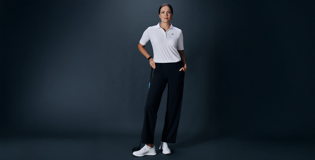 Eine Frau steht vor dunklem Hintergrund. Sie trägt eine dunkelbaue Golfhose und dazu ein weißes Golfpolo.