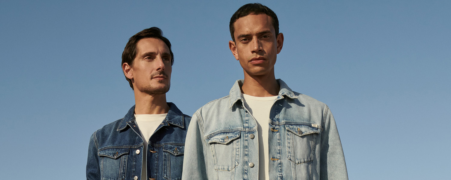 Zwei Männer in Jeansjacken, eine dunkelblau, die andere hellblau, vor klarem Himmel