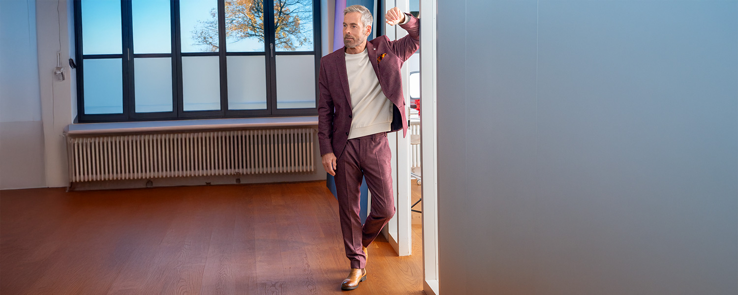 Mann in bordeauxrotem Anzug und beigem Pullover, stehend im Büro an Türrahmen.