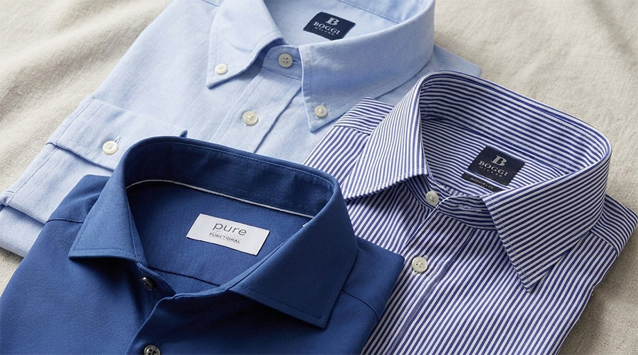 Drei gefaltete Business-Hemden in Hellblau, Dunkelblau und Blau-Weiß gestreift auf beigem Untergrund