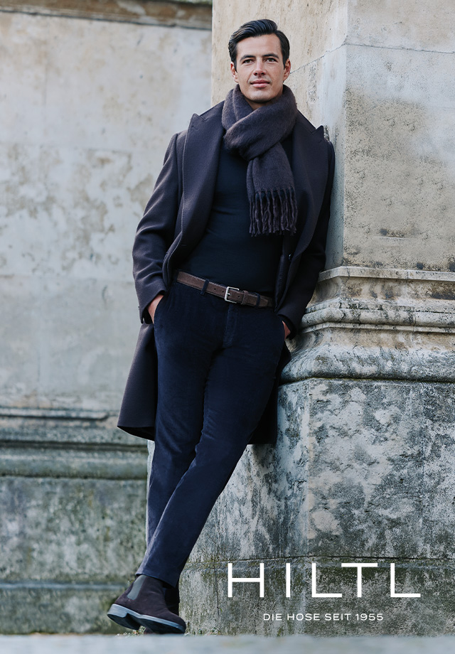 Mann in dunkelblauem Wollmantel mit grauem Schal, Business-Casual-Outfit, an Steinmauer gelehnt