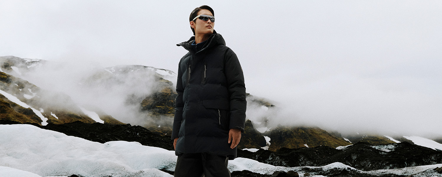 Mann in schwarzer Outdoor-Jacke mit Kapuze und Sonnenbrille in verschneiter Berglandschaft