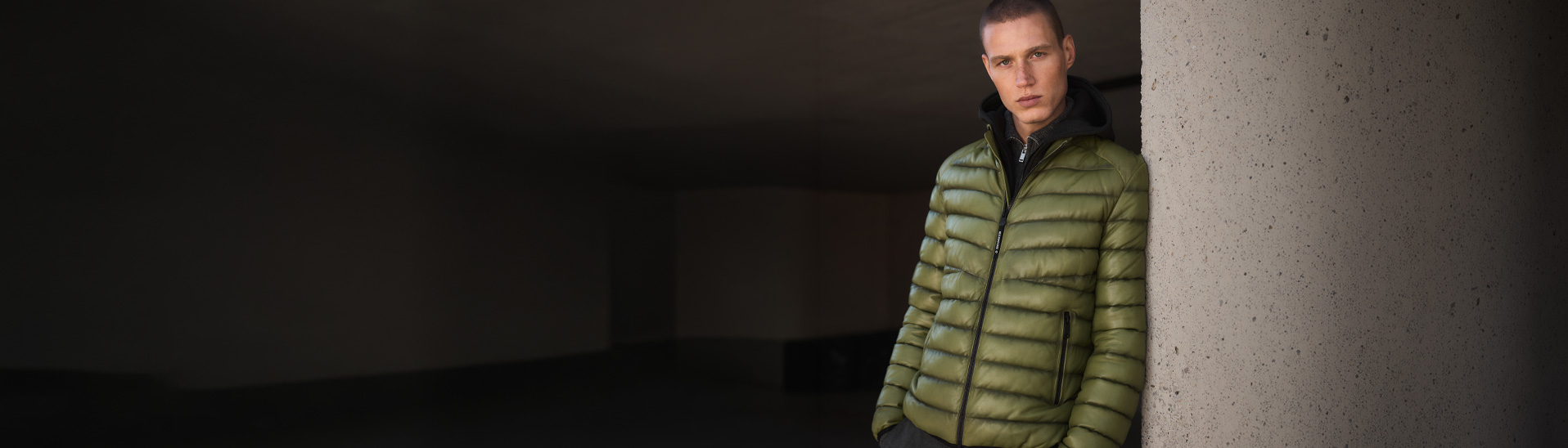 Mann in olivgrüner Steppjacke mit schwarzem Kapuzen-Futter, moderne Winterjacke vor Betonwand