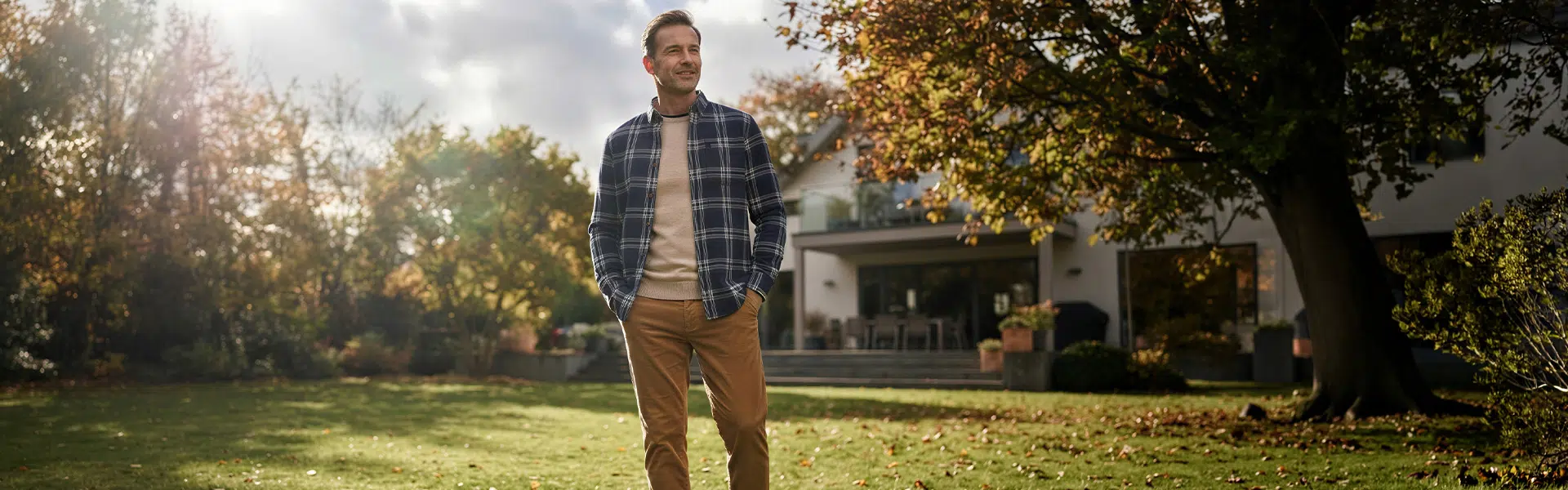Mann in kariertem Flanellhemd blau-grau über beigem Pullover und Chino-Hose im Garten