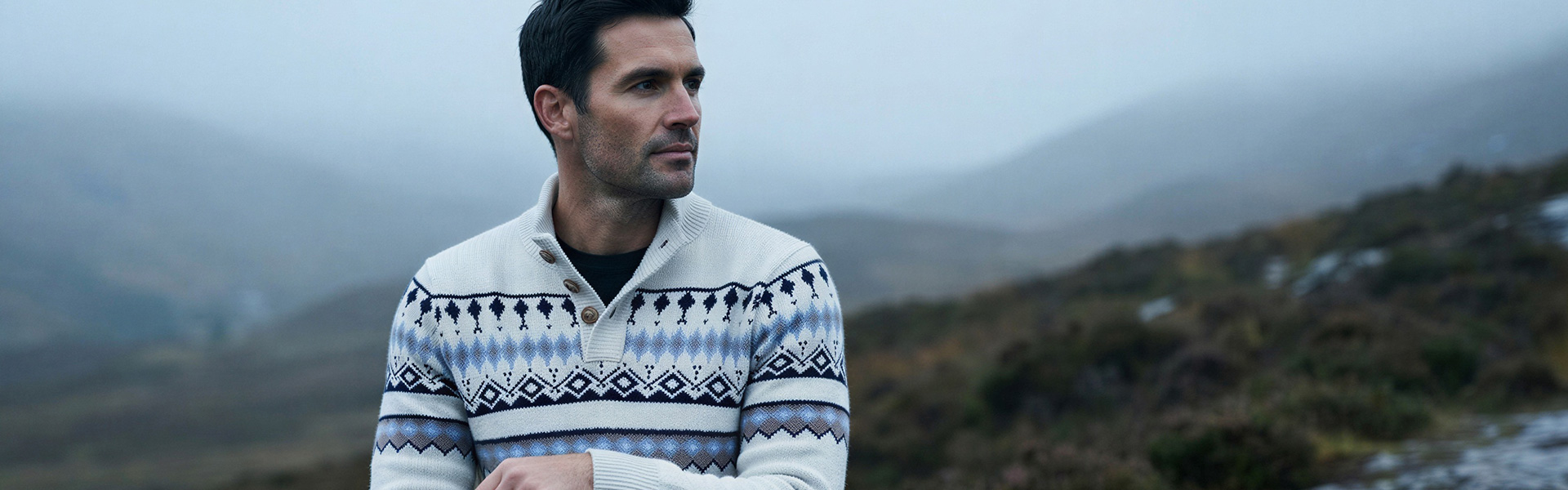 Mann in weißem Strickcardigan mit nordischem Muster in Blau und Grau, Berglandschaft im Hintergrund