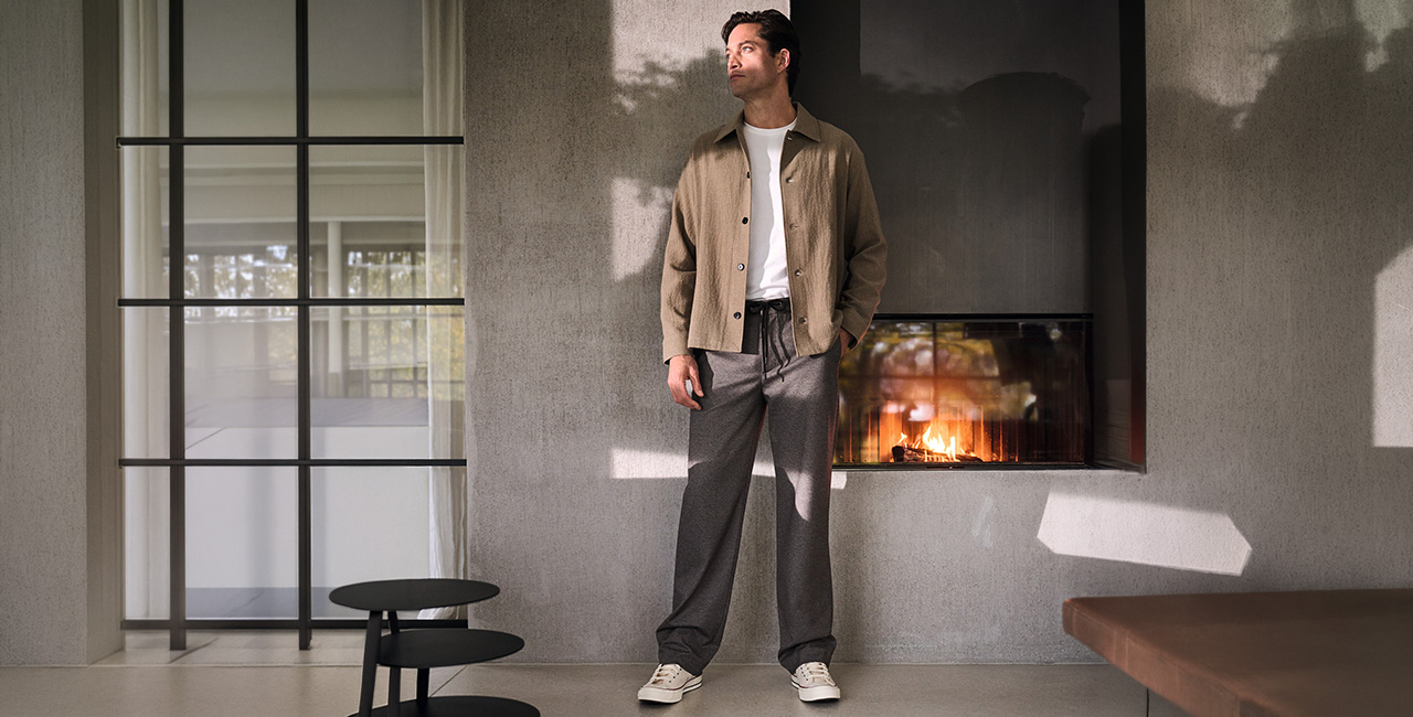 Mann in beiger Hemdjacke mit weißem T-Shirt und grauer Wide Fit Bundfalten-Hose, leger im modernen Wohnraum
