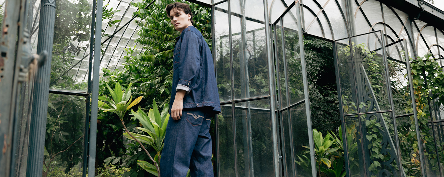Mann in blauer Jeansjacke und Jeans vor Glashaus mit Pflanzen