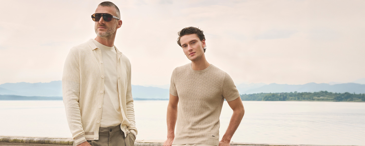 Herren in beigefarbenem Strickpullover und hellem Strickcardigan am Seeufer