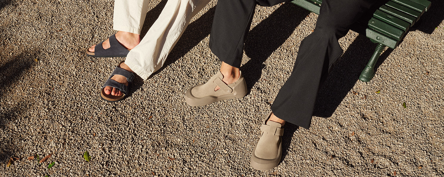Zwei Männerbeine in hellen und dunklen Hosen mit schwarzen Sandalen und beigefarbenen Slippern auf Kiesweg