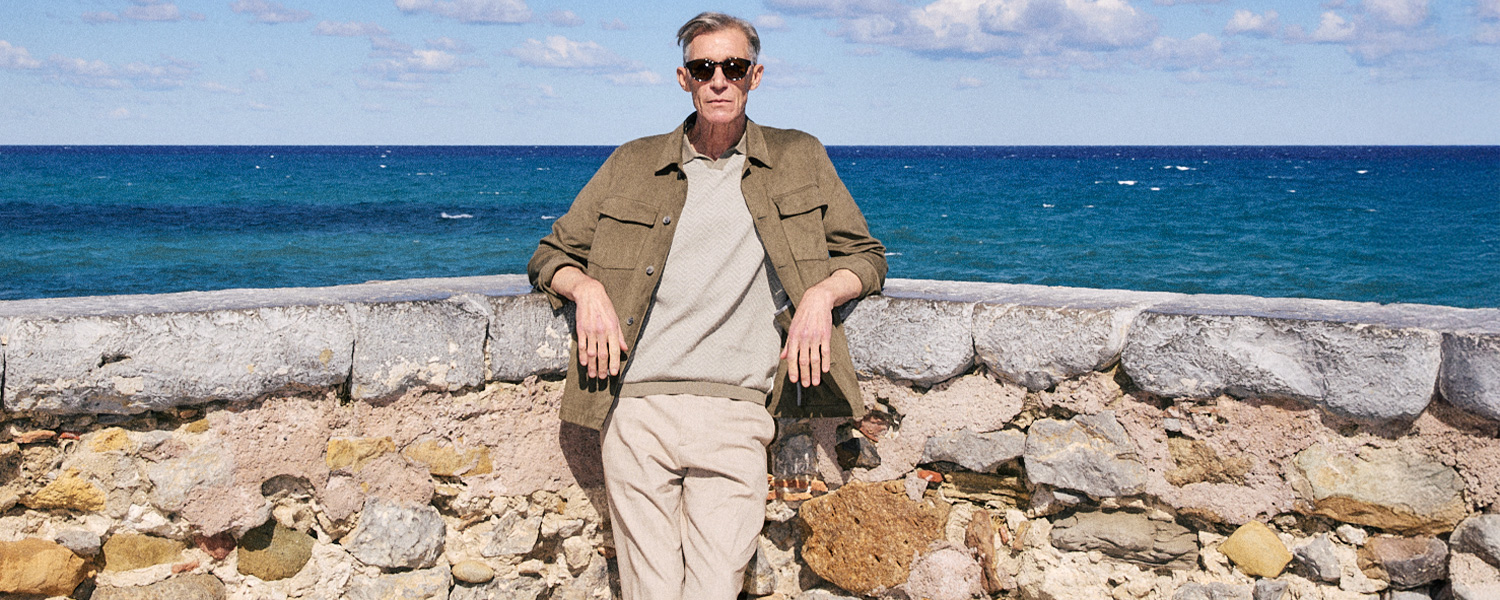 Mann in olivgrüner Jacke und beigem Pullover entspannt an Steinküste