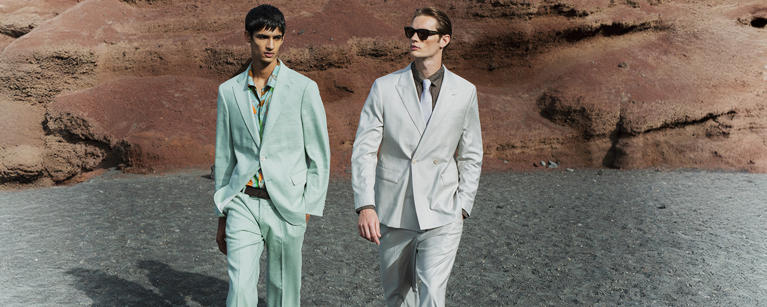 Zwei Männer in hellen Anzügen, einer mintgrün mit gemustertem Hemd, der andere beigefarben, spazieren draußen
