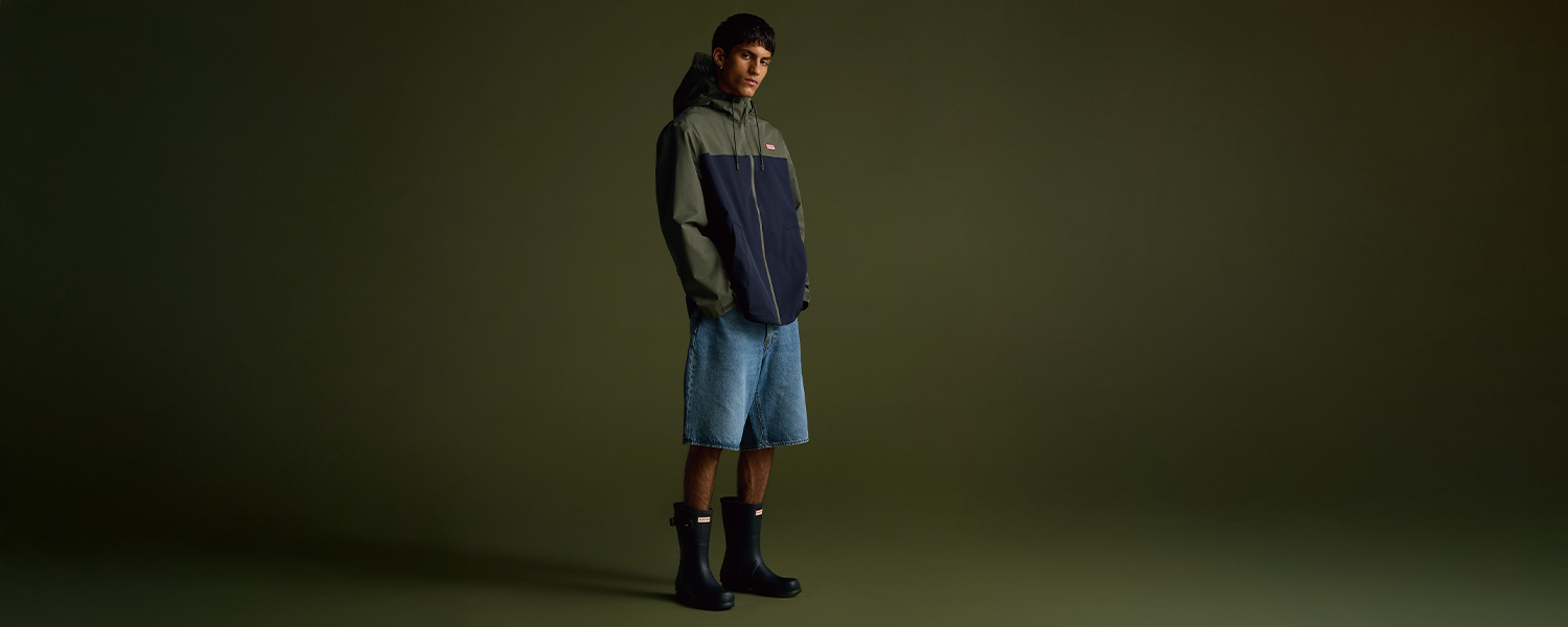 Mann in zweifarbiger Regenjacke, Jeansshorts und Gummistiefeln, leger präsentiert