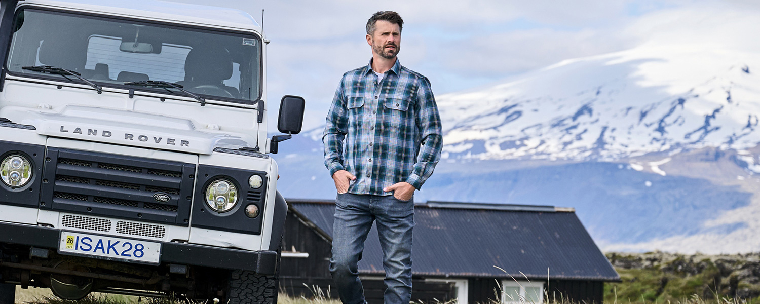 Mann in kariertem Freizeithemd aus Baumwolle neben Geländewagen, lässig in Berglandschaft