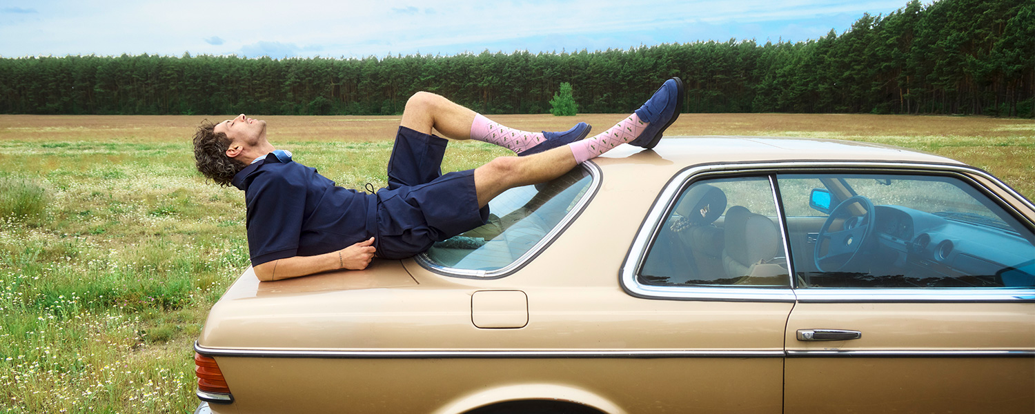 Mann in dunkelblauem Sommeranzug mit rosa Socken auf beigem Auto liegend im Freien