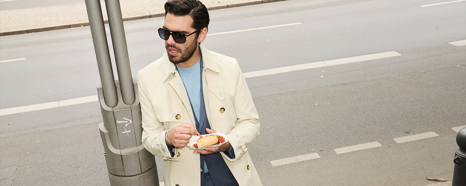 Mann in cremefarbenem Trenchcoat und blauem Pullover, lässig im Freien unterwegs