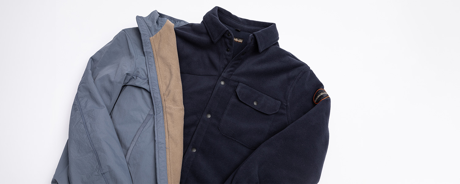 Auf dem Bild sind zwei gefaltete Fleecejacken zu sehen – eine in Dunkelblau mit Brusttasche und eine in Grau-Beige.