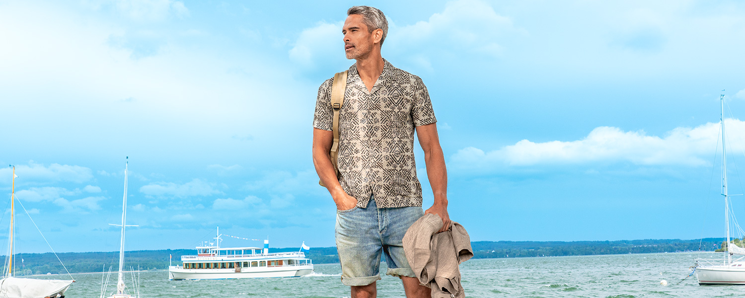 Ein Mann am Wasser trägt eine Jeans-Shorts, kombiniert mit gemustertem Hemd und Rucksack.