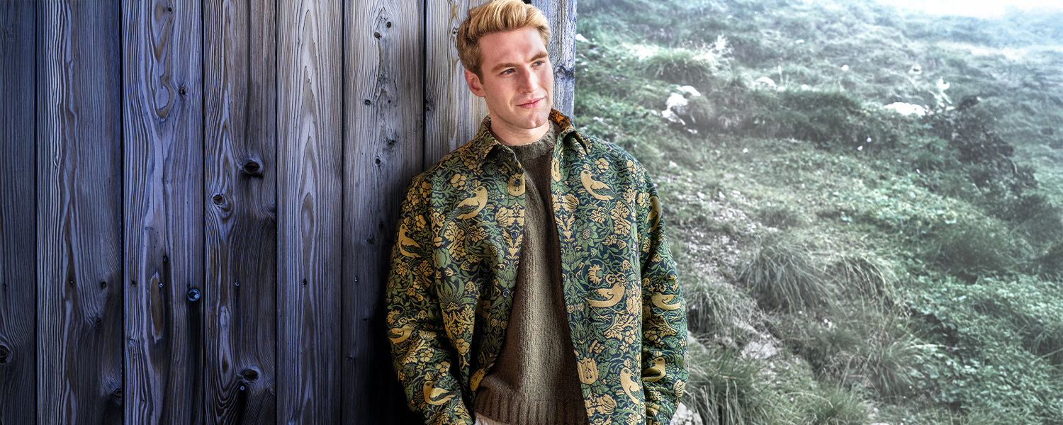 Herr in grauem Strickpullover mit nordischem Muster, lässig an Wand lehnend