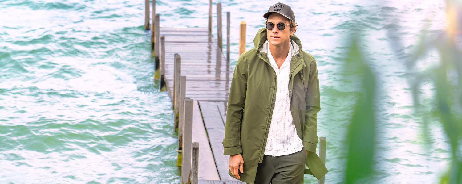 Grünes Cordhemd in extralanger Passform, getragen unter grauer Strickjacke mit Knopfleiste und Basecap im Outdoor-Setting.